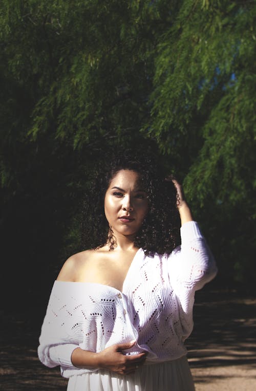 Young ethnic pensive woman with curly black hair wearing white outfit looking away in summer park