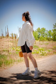 Woman in trendy attire standing outdoors in a park with handbag, embracing nature.