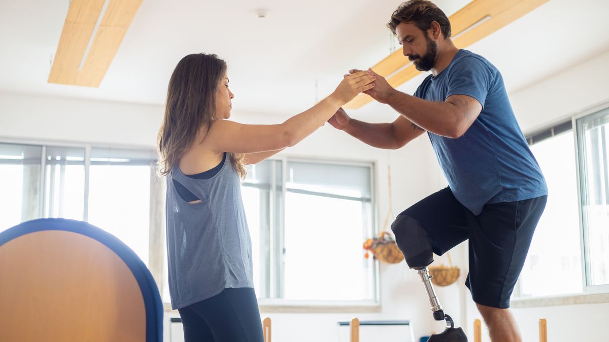 A man with a prosthetic leg exercises with a therapist in a well-lit gym environment.