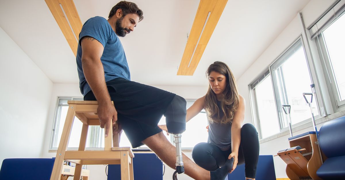 A trainer assists a man with a prosthetic leg during indoor physical therapy.