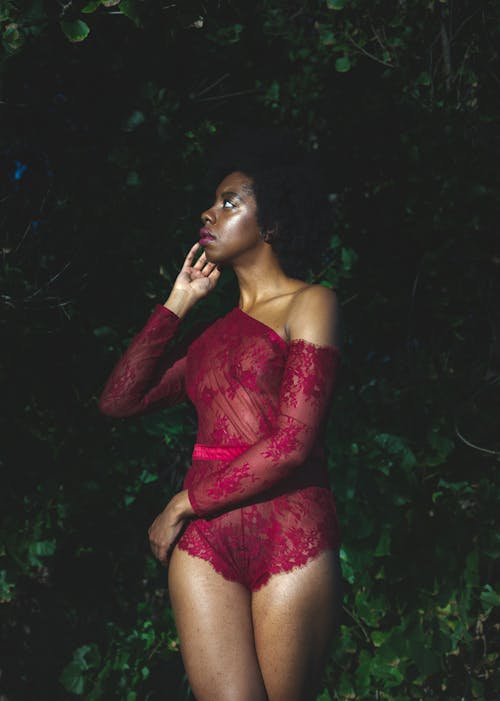 Young black female standing in silk dark red lace lingerie and looking away in night garden