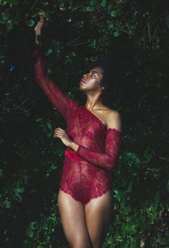 Young slender black female in red lace underwear standing in green garden at night