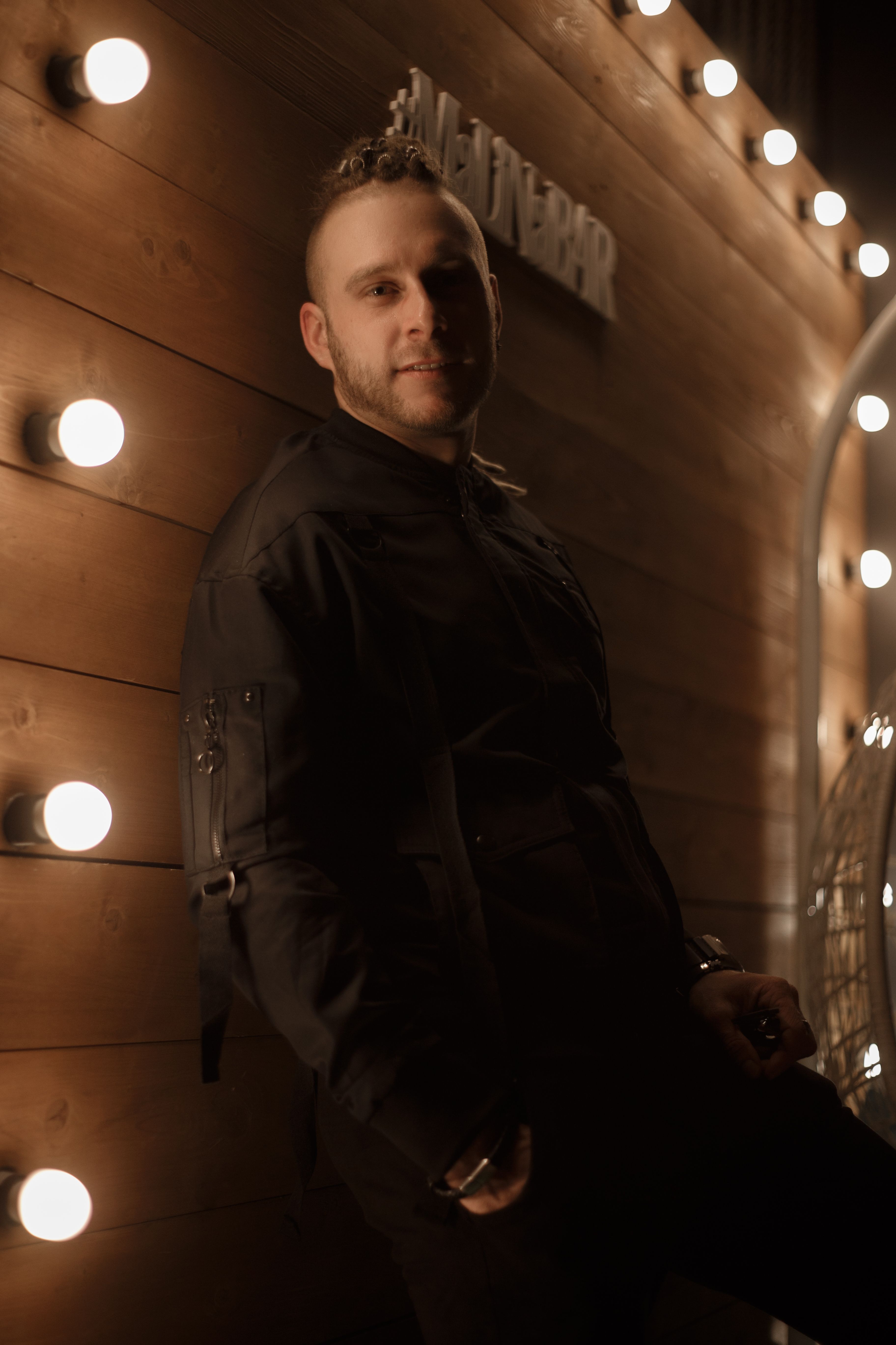 Young handsome cheerful male with beard looking at camera and smiling near wall with bright lamps in evening