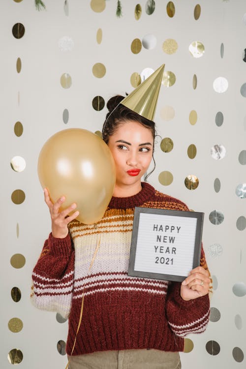 Content female wearing party hat standing on decorated white background with balloon and greeting inscription in hand while celebrating new year