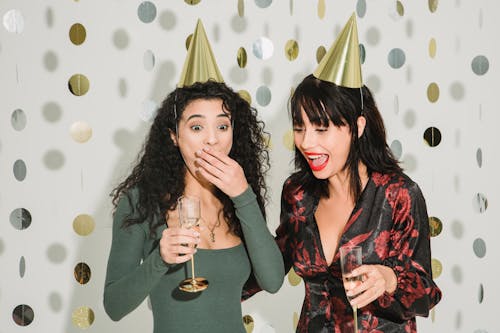 Free Surprised woman covering mouth while standing with glass of champagne near female friend in party hat during holiday celebration on white decorated background Stock Photo