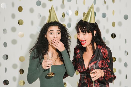 Surprised woman covering mouth while standing with glass of champagne near female friend in party hat during holiday celebration on white decorated background