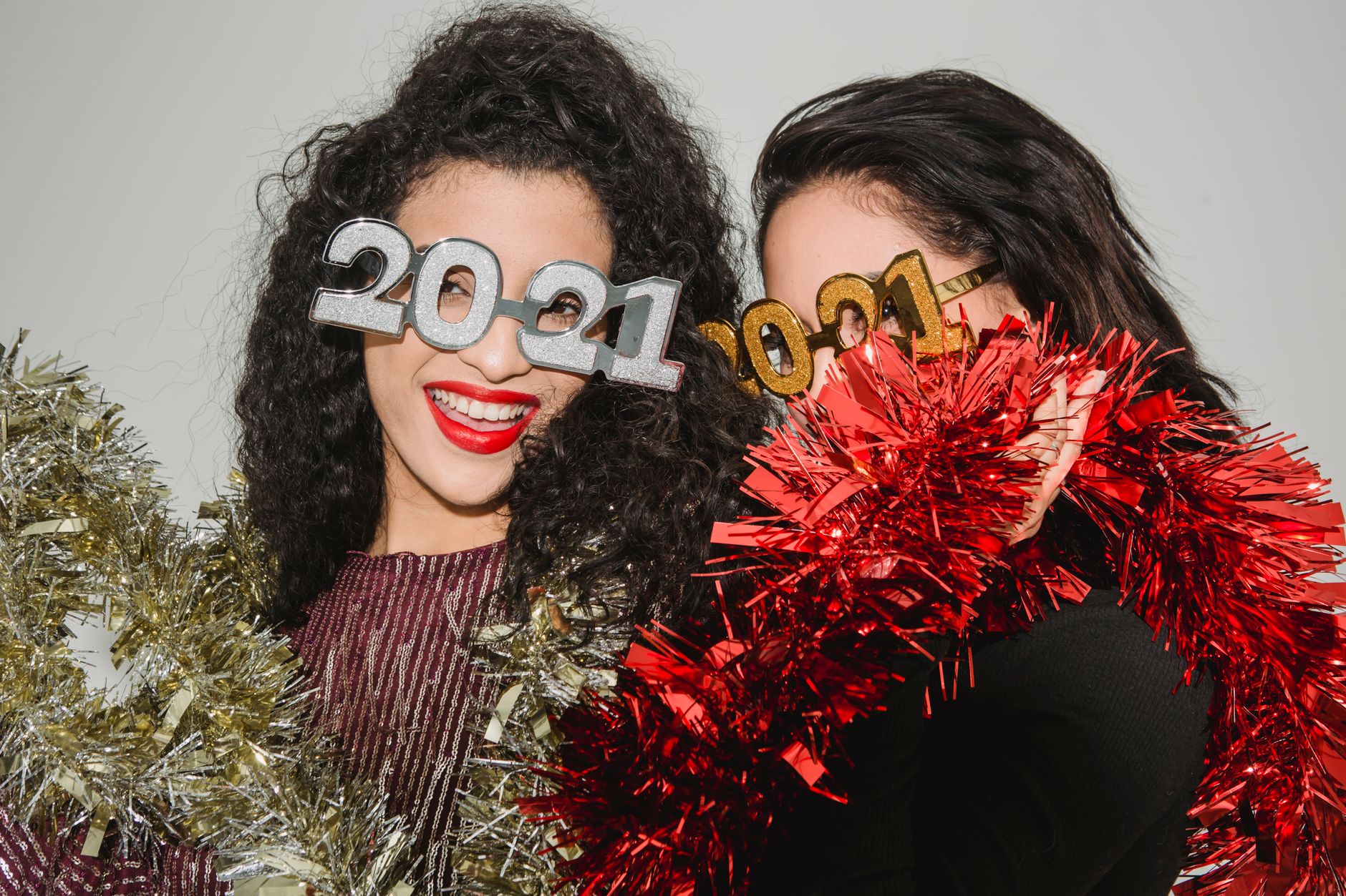 Women celebrating New Year's Eve with 2021 glasses and tinsel indoors, showcasing joyful expressions.