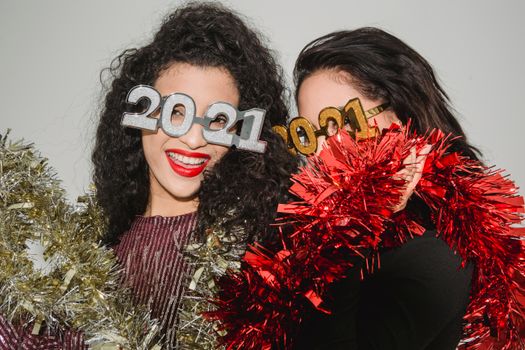 Women celebrating New Year's Eve with 2021 glasses and tinsel indoors, showcasing joyful expressions.