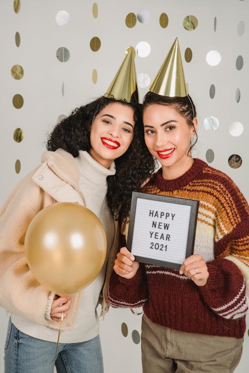 Smiling female wearing warm clothes looking at camera while standing with balloon and tablet with greeting inscription on white decorated background during party