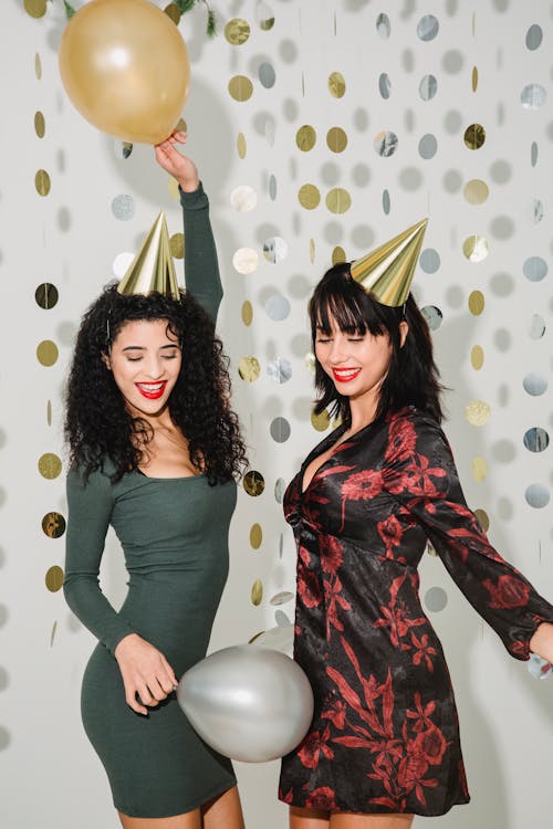 Free Happy female friends with helium balloons wearing party hats having fun while standing on white background with paper garland during holiday celebration Stock Photo