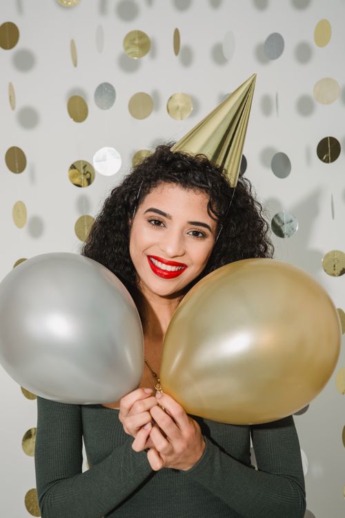 Free Cheerful female with dark hair wearing party hat looking at camera while standing with colorful balloons on white background with hanging decoration Stock Photo