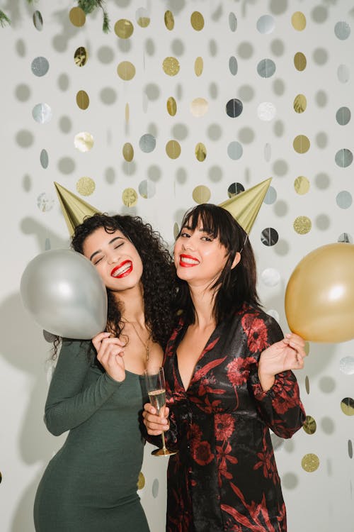 Delighted female friends wearing party hats standing with balloons and champagne glass  on white background with hanging circle dots