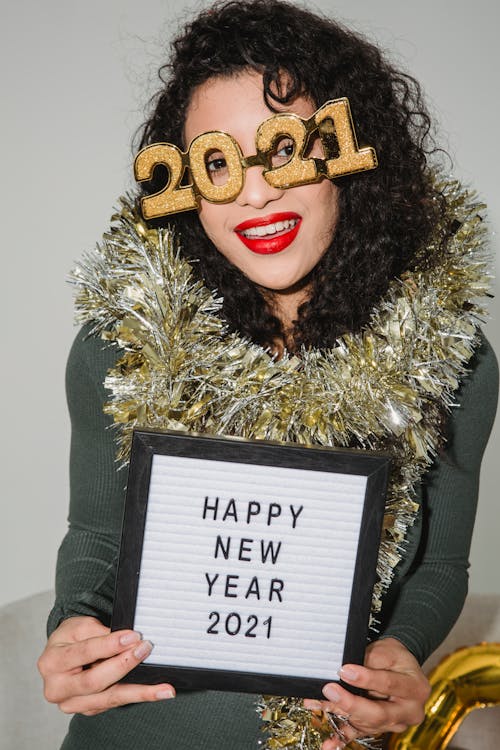 Positive female with tinsel wearing text glasses 2021 looking at camera while sitting on white background with greeting inscription on tablet in hands