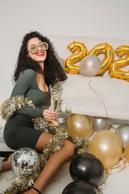 Delighted female wearing striped glasses and tinsel sitting with glass of champagne near balloon numbers in studio during holiday celebration