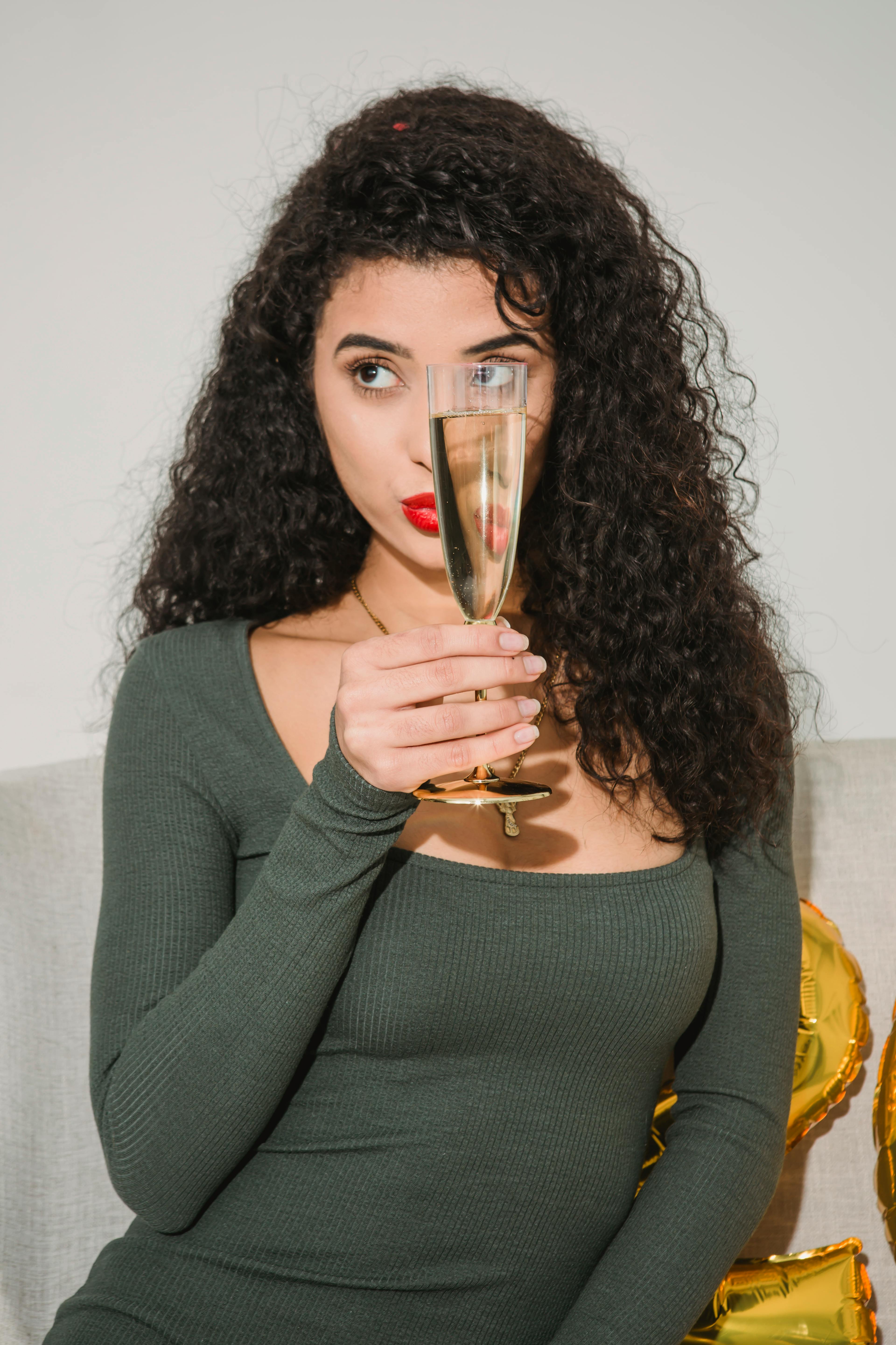 Content female with curly hair and red lips celebrating special event with glass of champagne and looking away