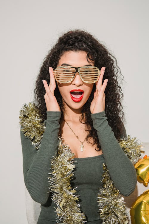 Young astonished female with red lips and shimmering golden tinsel touching striped glasses and looking at camera on white background