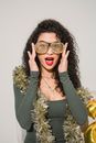Young astonished female with red lips and shimmering golden tinsel touching striped glasses and looking at camera on white background