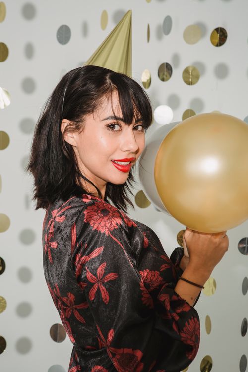 Free Side view of young female with red lips showing balloons and looking at camera among shimmering confetti at party Stock Photo