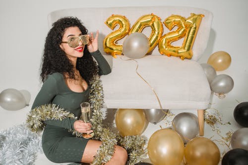 Cheerful young female with red lips and curly hair in striped glasses with glass of champagne among balloons