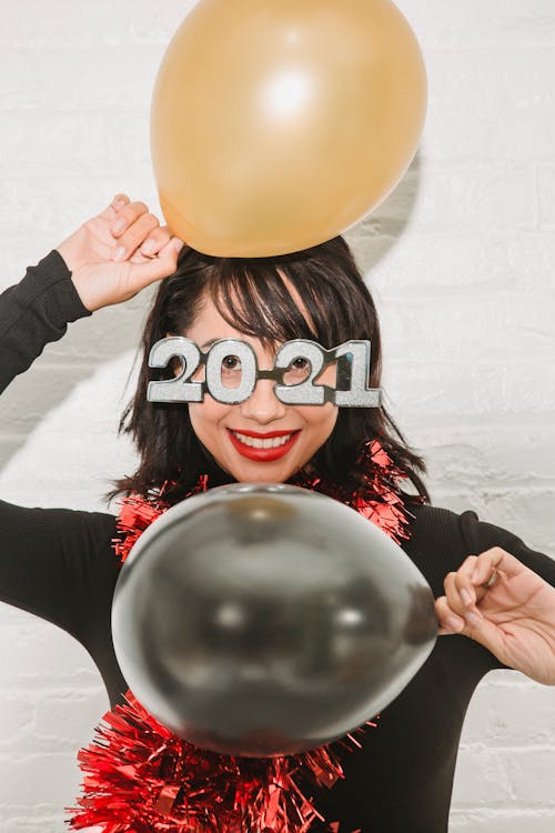 Cheerful young female in 2021 New Year glasses and tinsel looking at camera and demonstrating shiny balloons