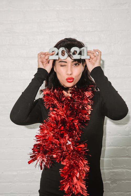 Young female in bright red sparkling tinsel sending air kiss and touching festive glasses near white brick wall
