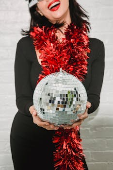 Chic woman in red tinsel with sparkling mirror ball, ready to celebrate indoors.