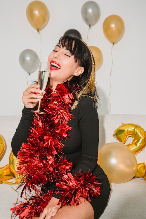 Cheerful female with mouth opened in tinsel preparing for drinking glass of champagne near silver and golden balloons
