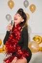 Cheerful female with mouth opened in tinsel preparing for drinking glass of champagne near silver and golden balloons