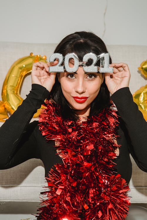 Young glad female in festive New Year eyeglasses and shimmering red tinsel looking at camera
