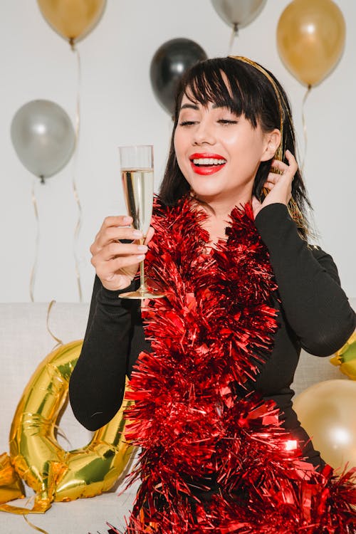 Happy young female with red lips and glass of champagne in shimmering tinsel smiling among balloons