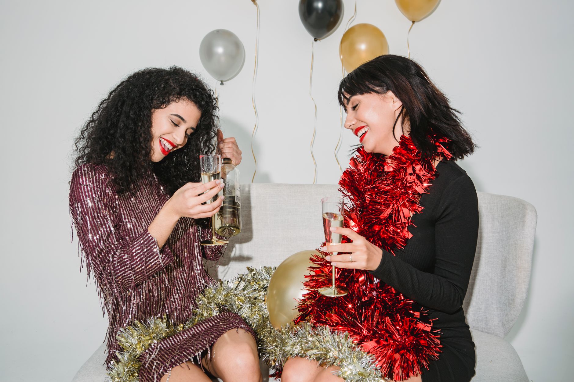 Cheerful young friends with red lips and dark hair entertaining and drinking champagne among balloons