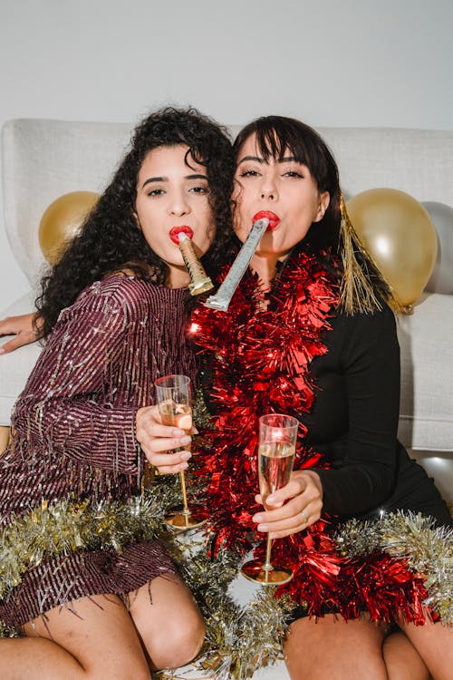 Cheerful young friends with red lips and shimmering tinsel looking at camera while entertaining at New Year party among balloons