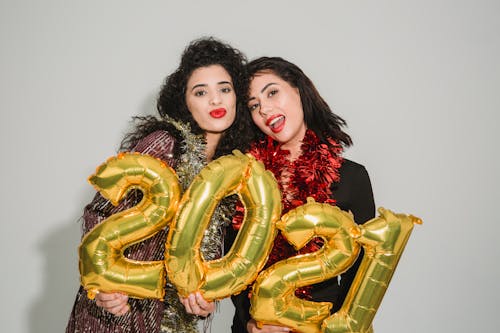 Happy women in festive clothes standing with golden balloons numbers 2021 against light wall during Christmas party