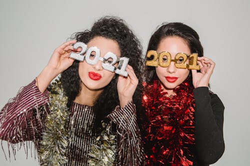 Positive friends in festive clothes and decorative eyeglasses of number 2021 standing near wall during New Year party and looking at camera