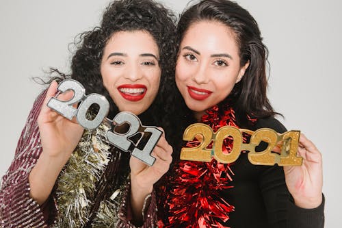 Cheerful women with dark hair in festive clothes with tinsels showing decorative numbers 2021 during New Year celebration against light wall