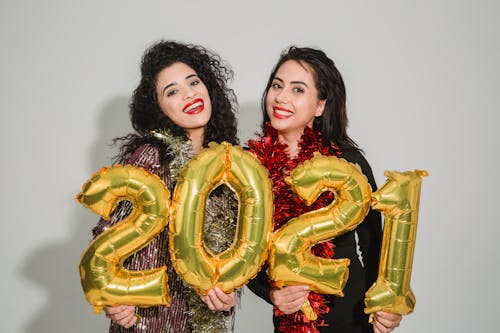Smiling women with dark hair in festive clothes standing with golden balloons numbers 2021 against light wall and looking at camera