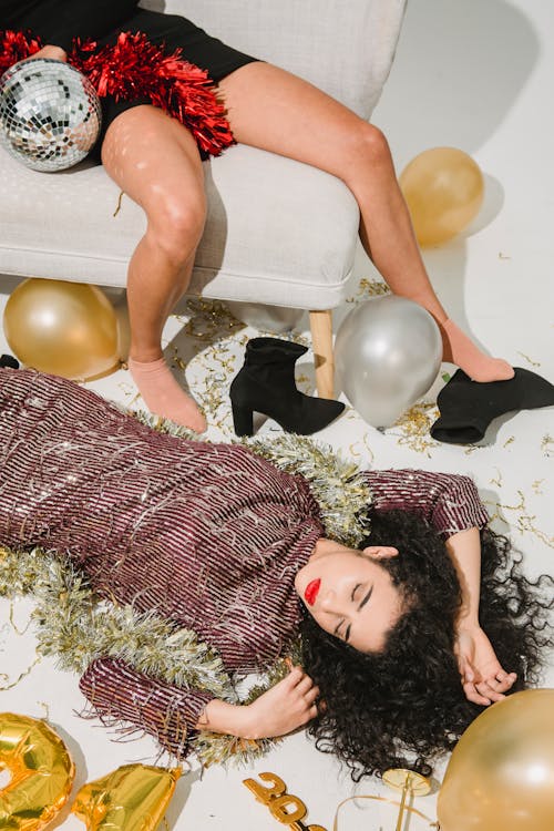 From above of drunk female in stylish dress sleeping on floor near friend lying on sofa in room with balloons and festive decorations after party