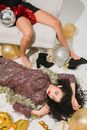 From above of drunk female in stylish dress sleeping on floor near friend lying on sofa in room with balloons and festive decorations after party