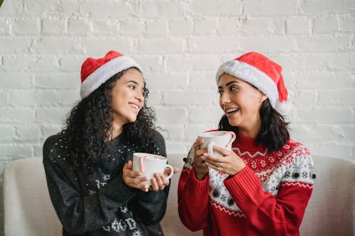 Cheerful women in sweaters and Christmas hats communicating and drinking cups of hot coffee against brick wall in room