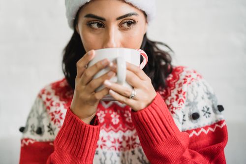 Free Young lady in Santa hat and sweater enjoying cup of cacao with candy cane in light room and looking away Stock Photo