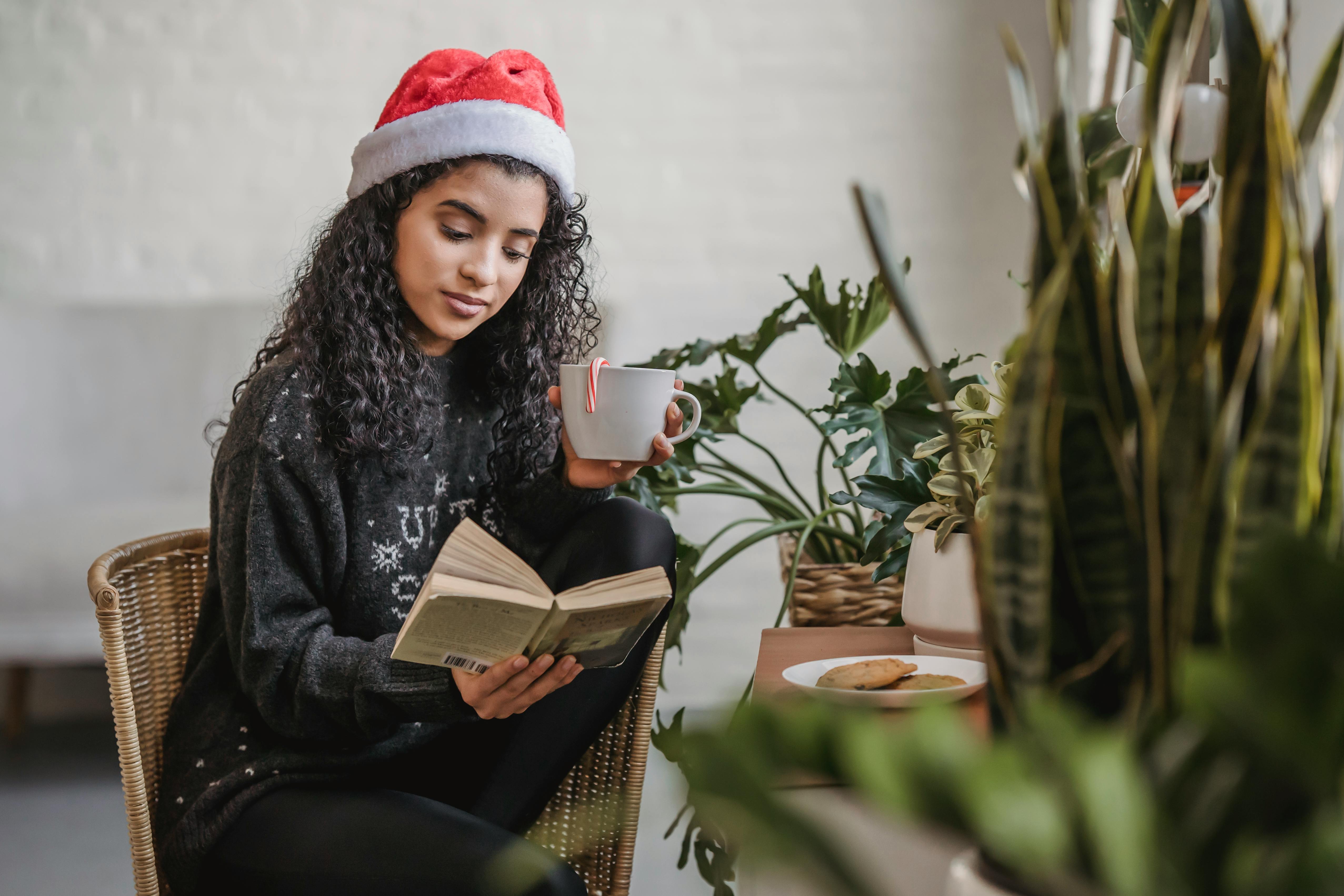 thoughtful woman in christmas hat reading book in cafe