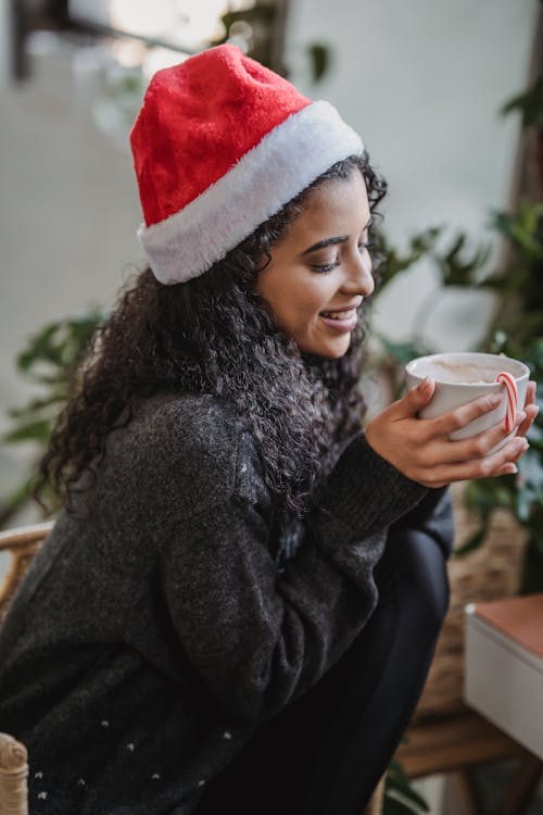 Side view of young ethnic female in warm clothes and Santa hat drinking hot coffee