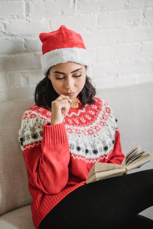Free Young female in warm Christmas sweater and Santa hat sitting on sofa and reading book Stock Photo