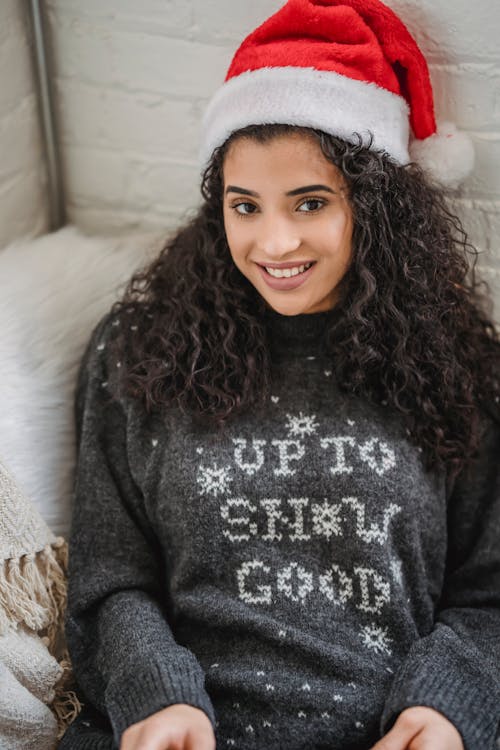 From above of cheerful young woman in warm clothes and hat smiling and sitting on armchair on Christmas