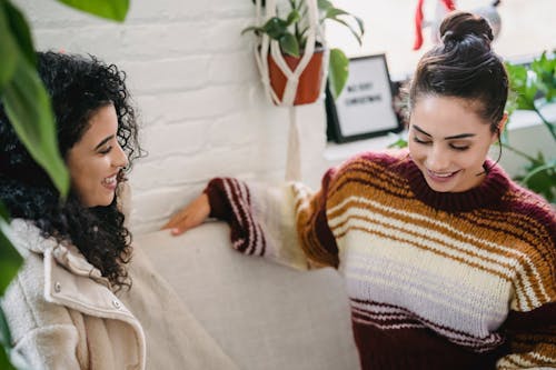 From above of happy young diverse females in casual clothes smiling and talking while sitting on comfortable couch in cozy living room