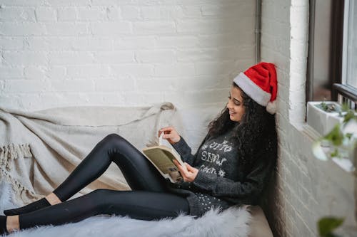 Side view of young female in casual clothes and Santa hat resting on sofa and reading book in modern living room on Christmas