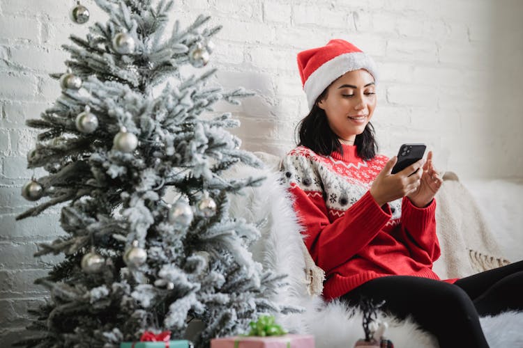 Cheerful Woman Using Smartphone Near Christmas Tree In Apartment