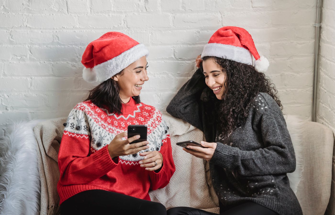 Two women wearing Santa hats and festive sweaters enjoying a cheerful conversation indoors during the holiday season.