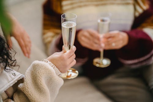Unrecognizable women in casual clothes celebrating holiday sitting on sofa with glasses of champagne