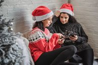 Cheerful women watching video on smartphone sitting on sofa near Christmas tree in room